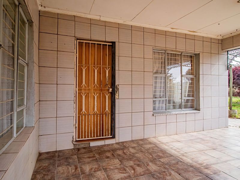 A covered patio area featuring tiled walls and flooring, equipped with a security gate and steel-framed windows overlooking the garden.