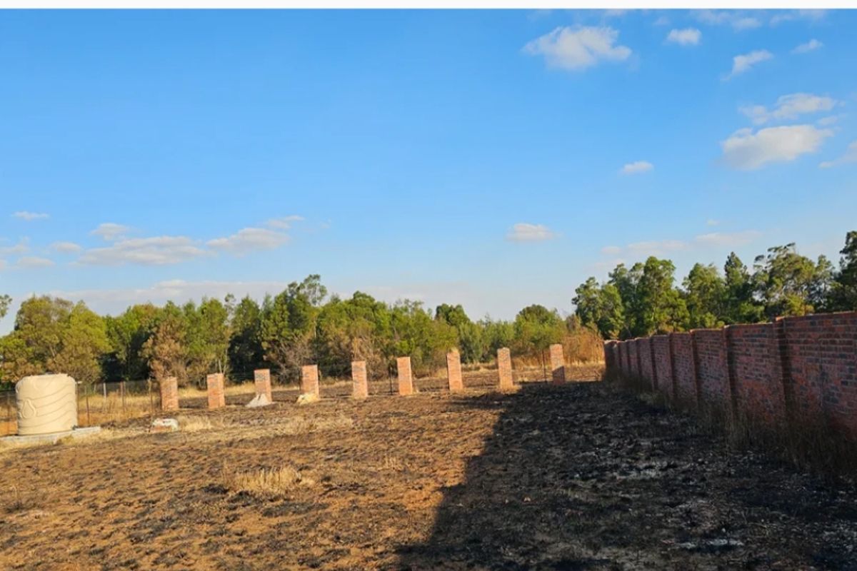 A vacant residential plot featuring a brick boundary wall, fence pillars, and a water storage tank on a cleared, scorched landscape.