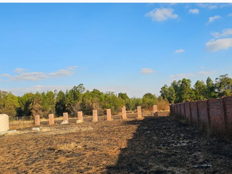 A vacant residential plot featuring a brick boundary wall, fence pillars, and a water storage tank on a cleared, scorched landscape.