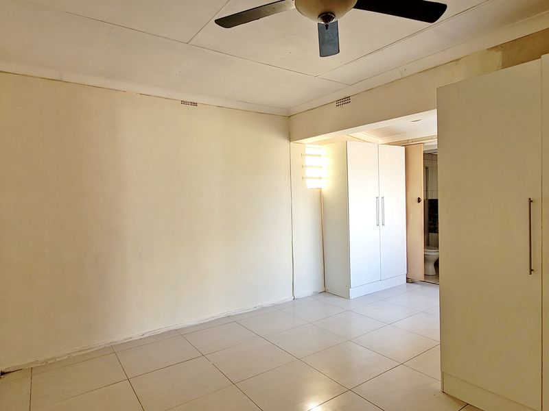 A spacious, empty bedroom featuring neutral tiled flooring, built-in cupboards, and a ceiling fan, offering a blank canvas for personalization.