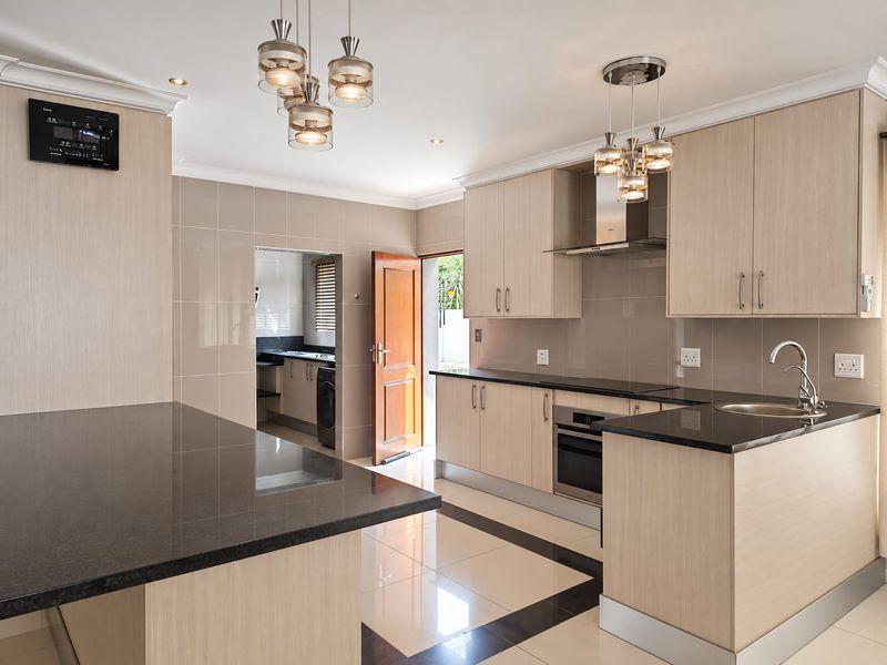 Modern kitchen featuring light wood cabinetry, dark granite countertops, integrated appliances, and tiled flooring with a seamless transition to the scullery area.