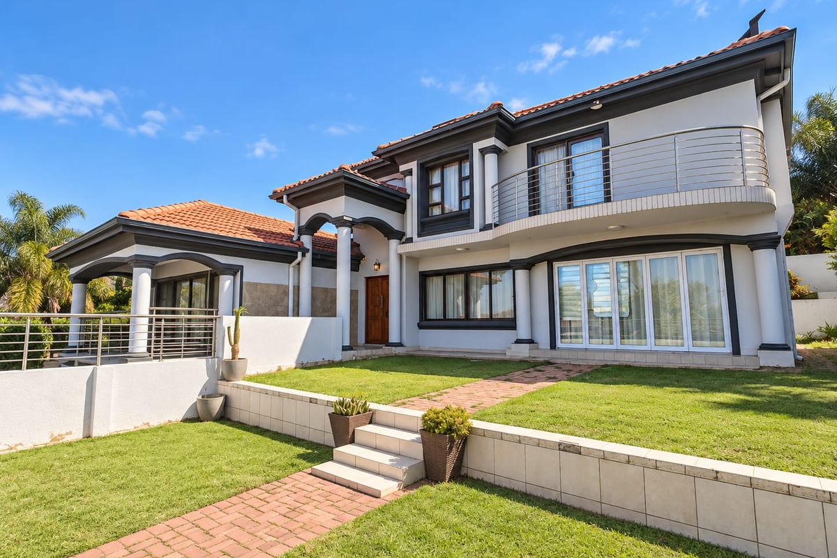 A well-maintained rear exterior view featuring a spacious lawn, brick pathways, a covered patio, and an upper-level balcony with modern stainless steel railings.