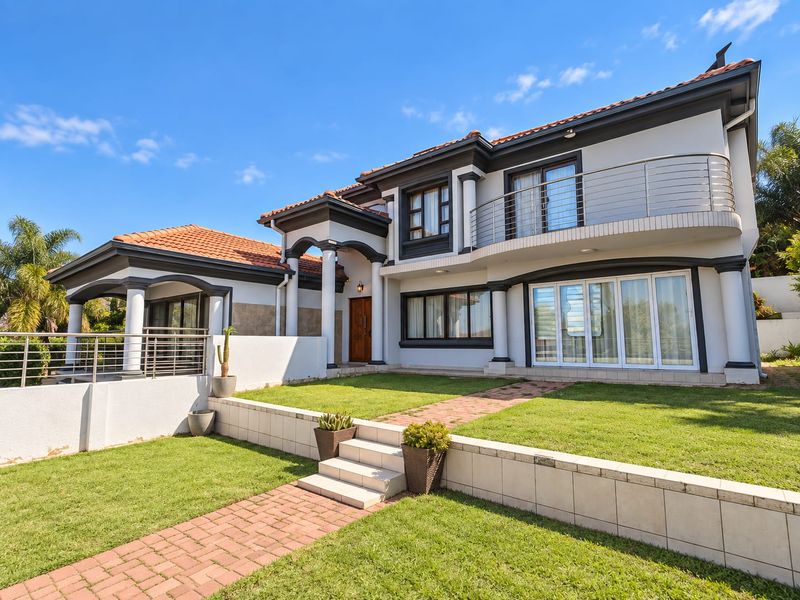 A well-maintained rear exterior view featuring a spacious lawn, brick pathways, a covered patio, and an upper-level balcony with modern stainless steel railings.