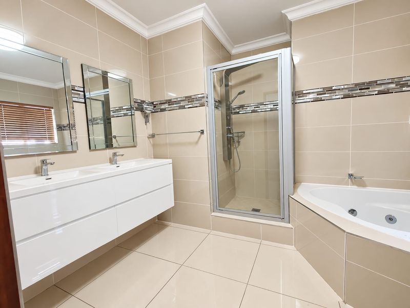 Modern bathroom featuring a double vanity, glass-enclosed shower, and a corner spa bath with neutral wall and floor tiling.