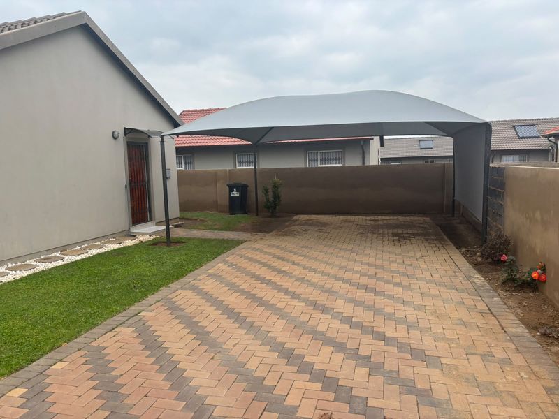 Front exterior view featuring a paved driveway, a modern shade port, a well-maintained lawn, and a secure boundary wall.