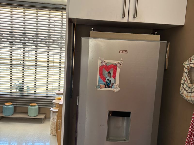 A functional kitchen area featuring built-in cabinetry, a countertop, and a built-in washing machine, with natural light provided by a window with venetian blinds.