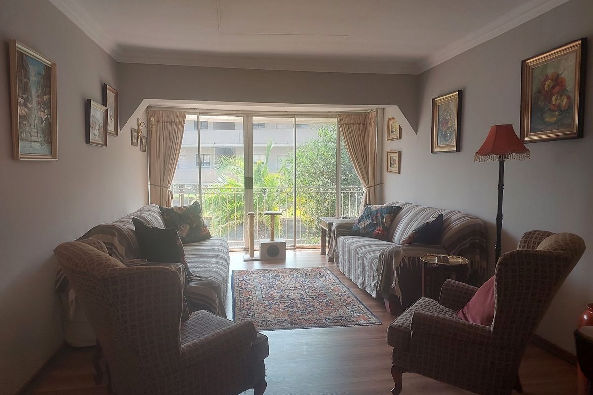 A comfortable living room featuring laminate flooring, cove ceiling details, and a large sliding glass door providing access to a balcony with garden views.