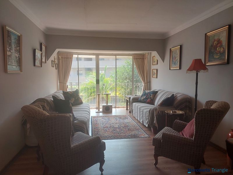 A comfortable living room featuring laminate flooring, cove ceiling details, and a large sliding glass door providing access to a balcony with garden views.