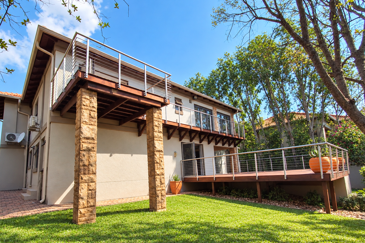 Rear view of the property featuring a raised timber deck with stainless steel balustrades, stone-clad columns, and a well-maintained garden.