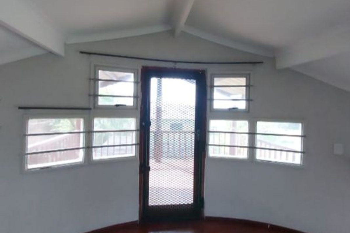 An empty living area featuring exposed ceiling beams, multiple casement windows with security bars, and a security screen door leading to an outdoor space.