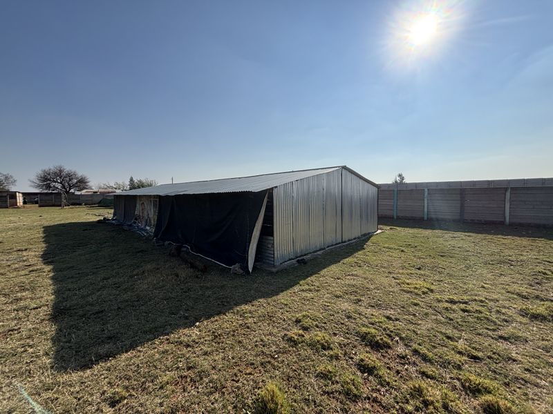 A view of a corrugated metal outbuilding or shed structure situated on a large plot, enclosed by a precast concrete boundary wall.