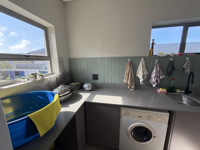 A functional laundry room featuring modern grey cabinetry, stone countertops, a stainless steel sink, and a tiled splashback with natural light.