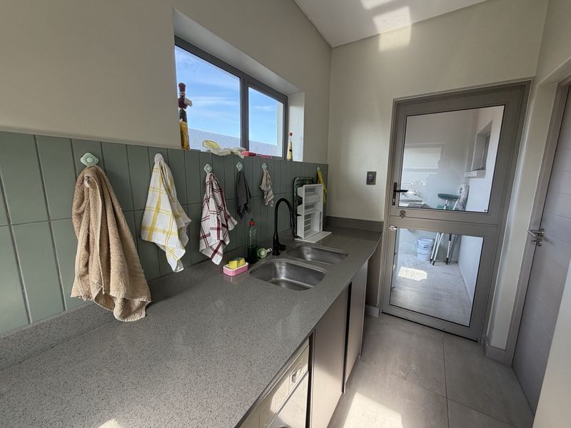 A functional scullery featuring a double stainless steel sink, stone-look countertops, tiled splashback, and direct access to the exterior via a glass-panelled door.
