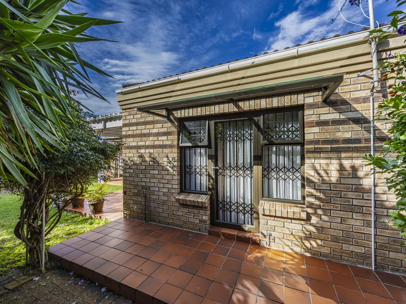 A rear exterior view of the townhouse featuring a brick facade, a tiled patio area, and a secure entrance door with a metal security gate.