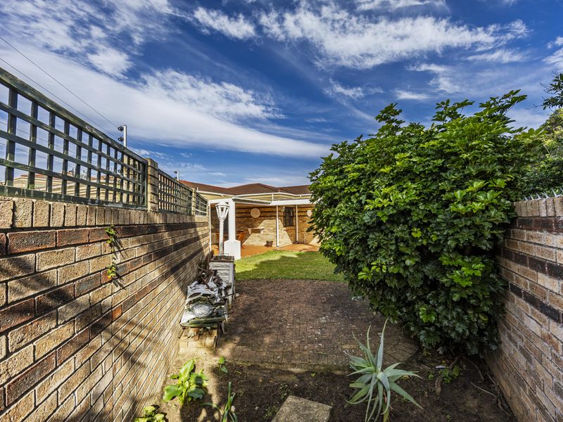 A rear exterior view of the townhouse featuring a brick boundary wall, paved pathway, small lawn area, and a covered patio space.