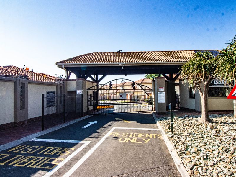 The secure entrance gate of the residential cluster complex, featuring controlled access, paved driveway, and perimeter security fencing.