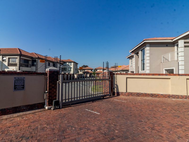 Entrance view of the cluster complex featuring a secure automated gate, brick perimeter walling with electric fencing, and a paved driveway.