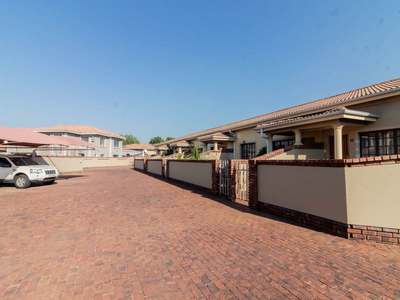 A view of the cluster complex entrance featuring a paved driveway, covered carports, and a row of residential units with tiled roofs and brick boundary walls.