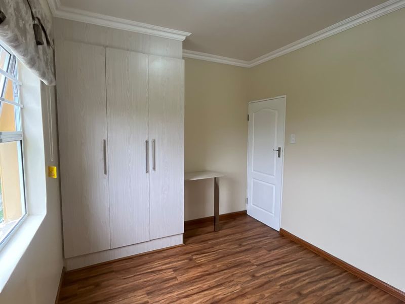 A bright, empty bedroom featuring modern laminate flooring, built-in cupboards with a small integrated desk, and crisp white walls with ceiling cornicing.