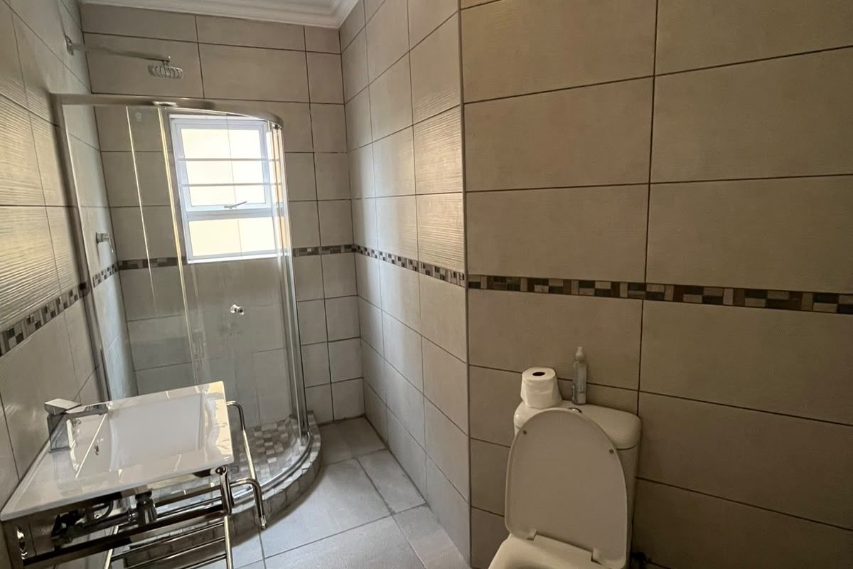 A functional bathroom featuring a corner shower enclosure, wall-mounted vanity basin, and toilet with neutral floor-to-ceiling wall tiling.