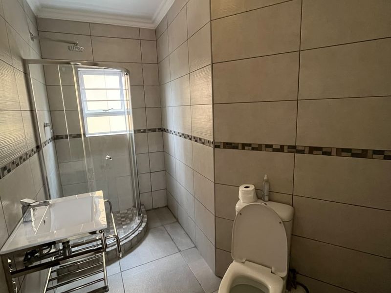 A functional bathroom featuring a corner shower enclosure, wall-mounted vanity basin, and toilet with neutral floor-to-ceiling wall tiling.