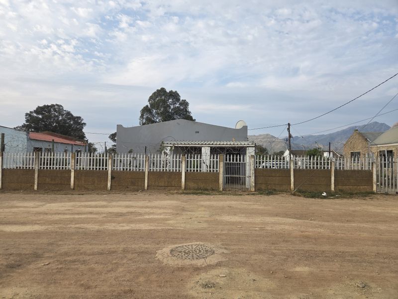 Front exterior view of a residential property featuring a perimeter wall with steel palisade fencing and razor wire, situated along an unpaved road.