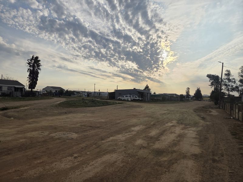 A wide-angle view of a residential street with unpaved road surfaces and surrounding neighborhood properties under a cloudy sky.