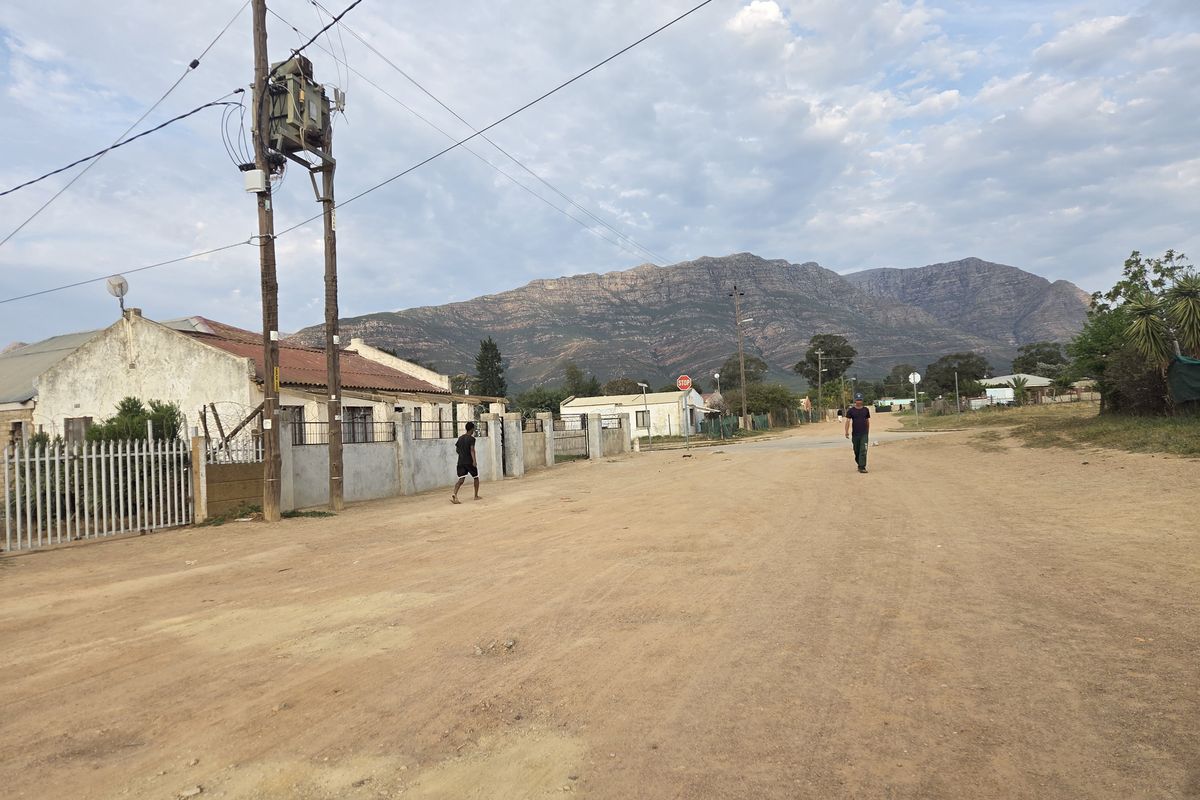 A street view showing the unpaved road approach to the property, featuring perimeter fencing and mountain views in the background.