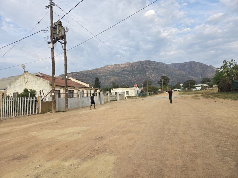 A street view showing the unpaved road approach to the property, featuring perimeter fencing and mountain views in the background.