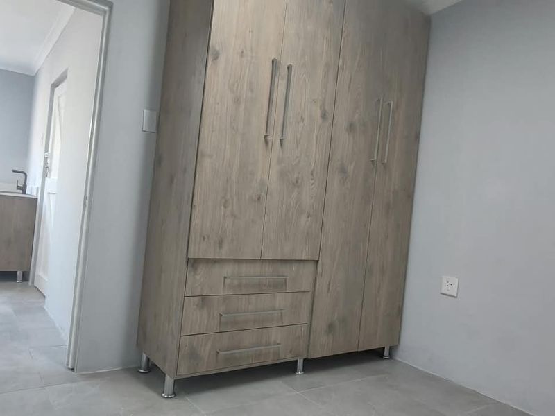A clean, modern bedroom featuring light wood-finish built-in wardrobes and neutral tiled flooring, with an adjacent view into a bathroom area.