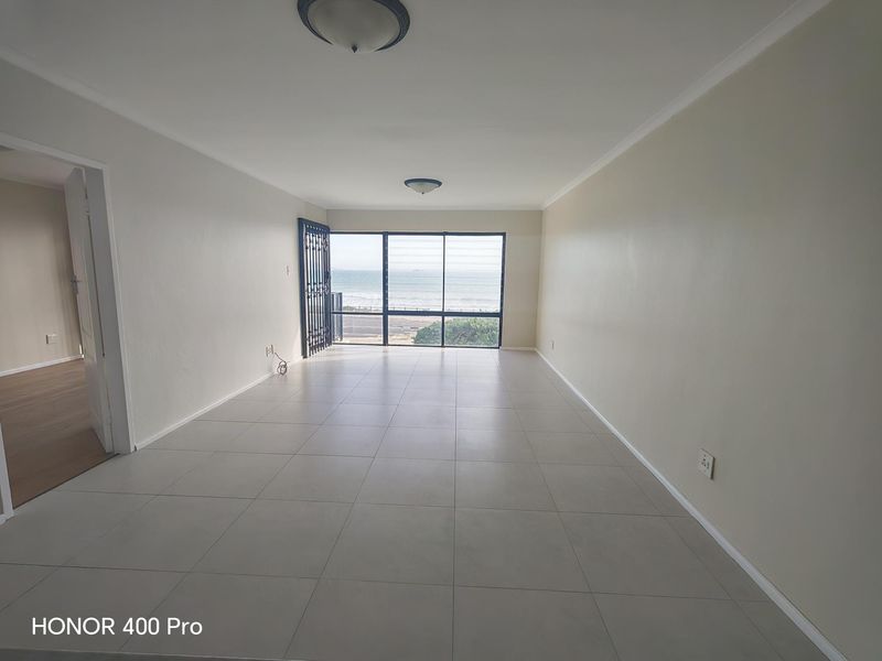 A spacious, empty living room featuring neutral tiled flooring, ceiling lighting, and a large window offering a direct ocean view.