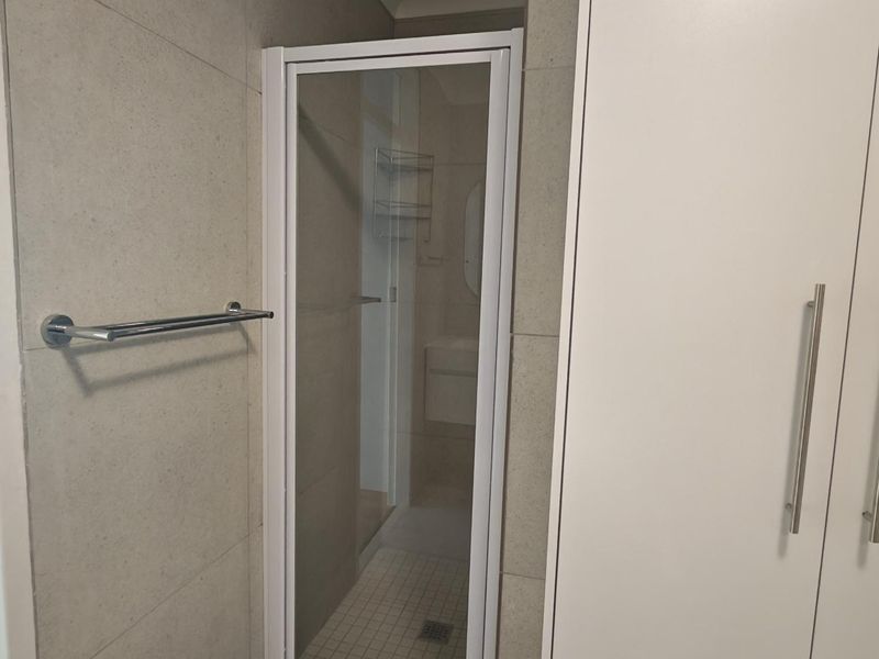 A clean, modern bathroom featuring a glass-enclosed shower, neutral wall tiling, and a built-in storage cupboard.