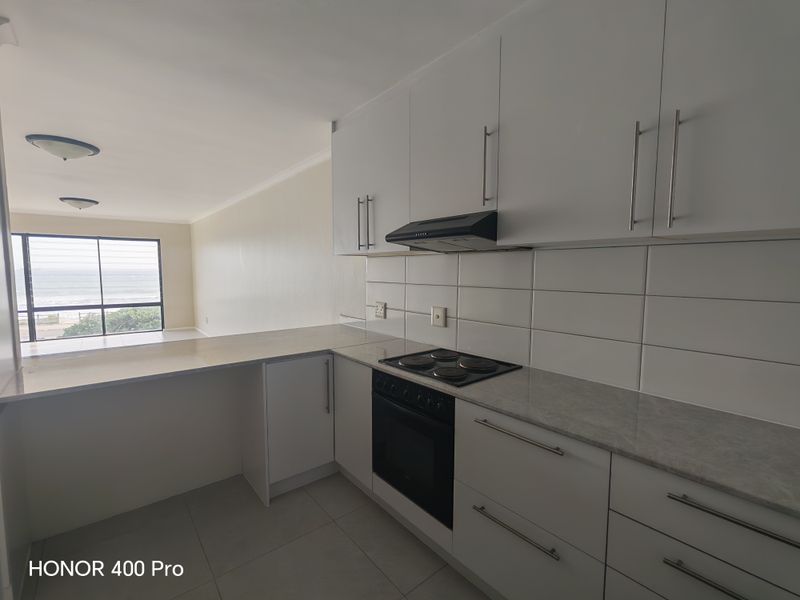 Modern open-plan kitchen featuring white cabinetry, stone-look countertops, and integrated appliances, flowing into a bright living area with ocean views.