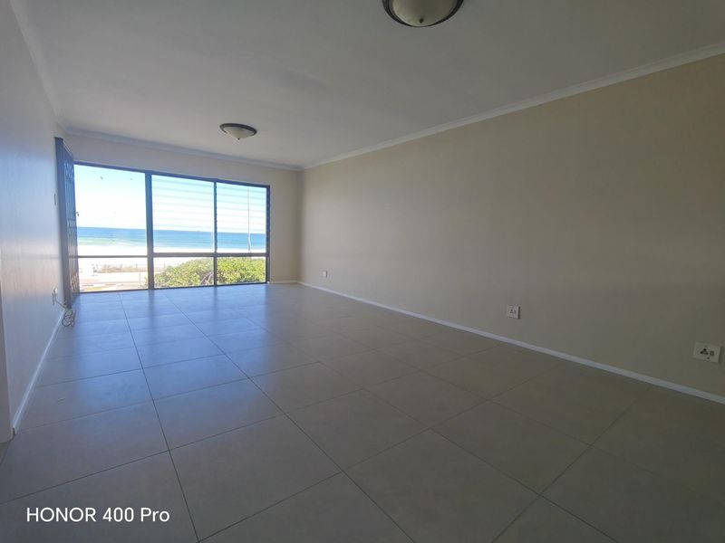 Spacious, empty living area featuring large windows with ocean views and neutral tiled flooring, offering a bright and versatile blank canvas.
