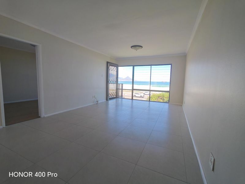 A bright, empty living room featuring neutral tiled flooring and a large sliding glass door with a security gate, offering scenic coastal views.
