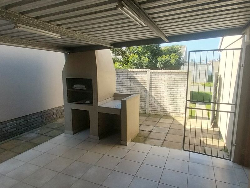 Covered outdoor patio area featuring a built-in braai, tiled flooring, and direct access to the rear garden through a security gate.