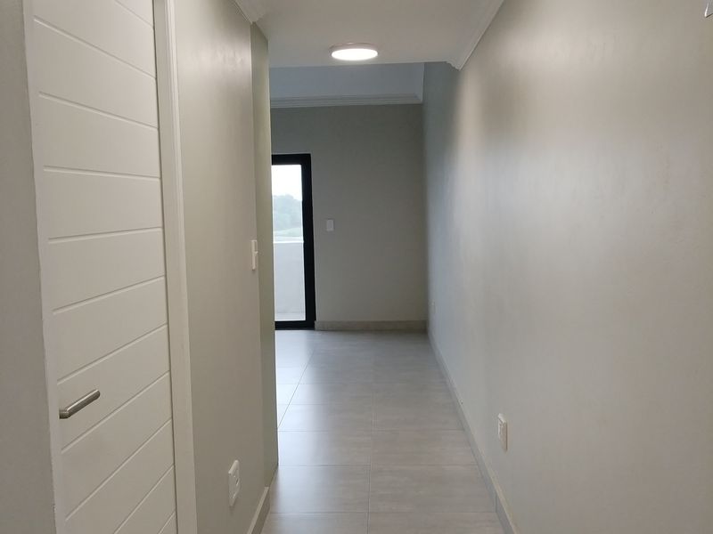 A clean, modern hallway featuring neutral wall tones, tiled flooring, and recessed ceiling lighting, leading towards an exterior doorway.