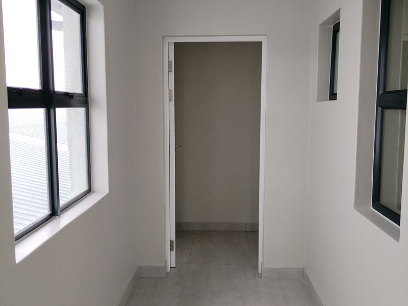 A bright, modern hallway featuring neutral wall tones, tiled flooring, and dark aluminium window frames, leading into an adjacent room.