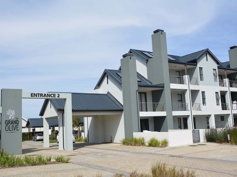 The front entrance of The Grand Olive apartment complex, featuring modern architectural design, paved driveway, and covered access.