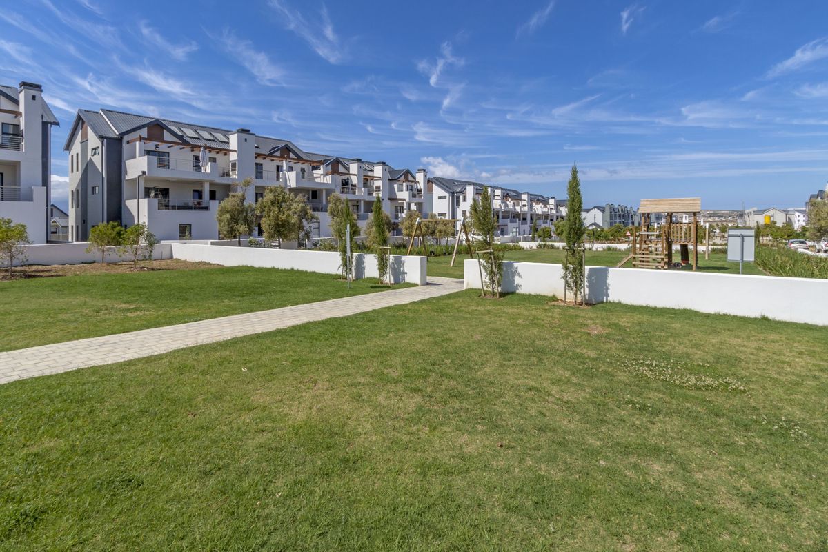 A well-maintained communal garden area featuring a paved walkway, manicured lawn, and playground equipment, set against the backdrop of modern apartment buildings.