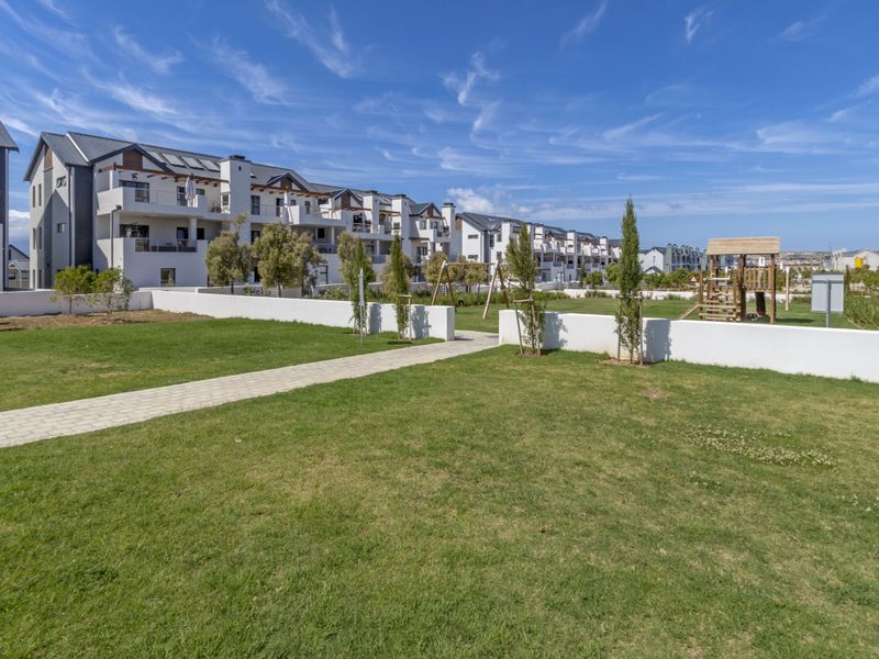 A well-maintained communal garden area featuring a paved walkway, manicured lawn, and playground equipment, set against the backdrop of modern apartment buildings.