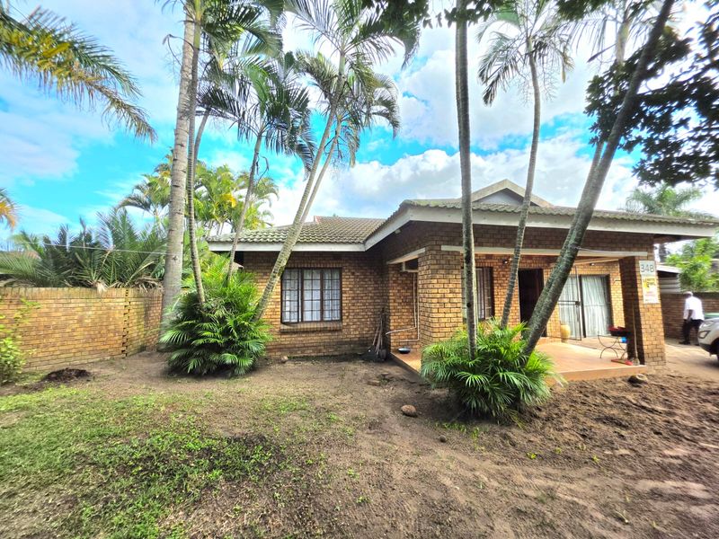 Front view of a face brick house featuring a covered patio, established palm trees, and a paved driveway area.