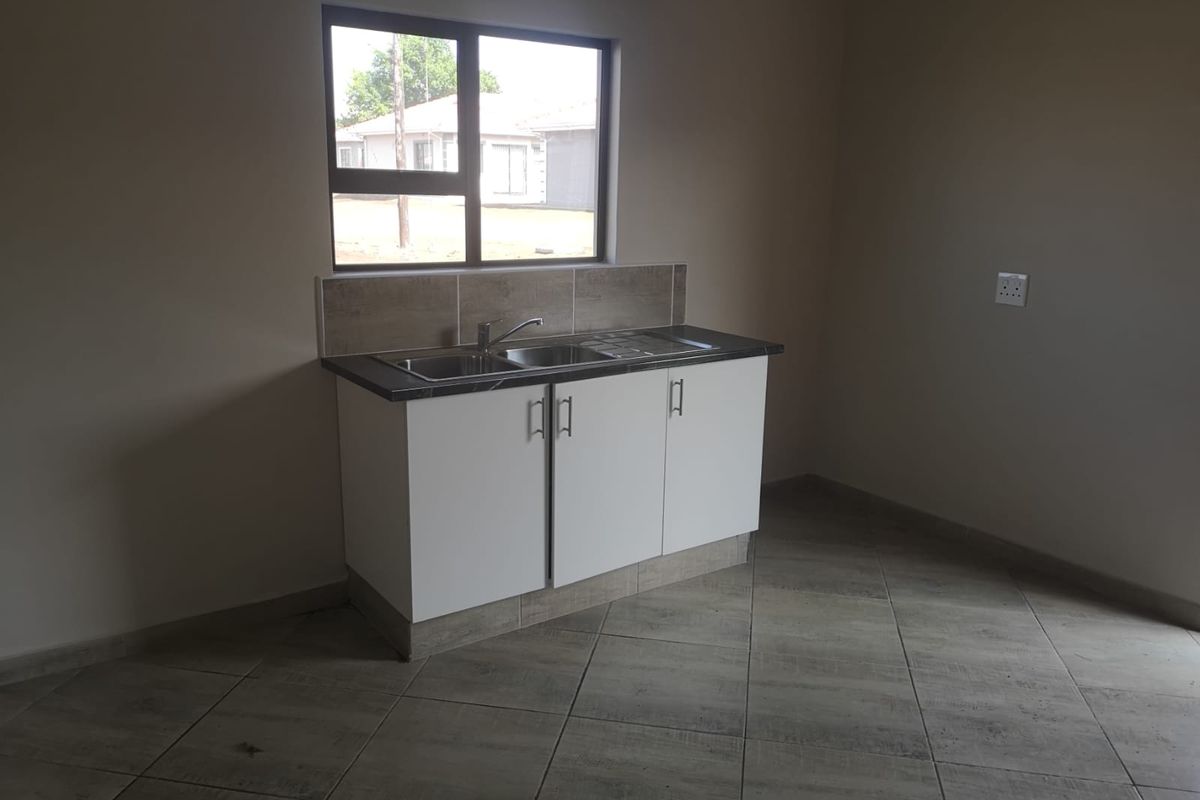 A compact, empty kitchen area featuring white cabinetry, a stainless steel sink, tiled splashback, and neutral floor tiles.