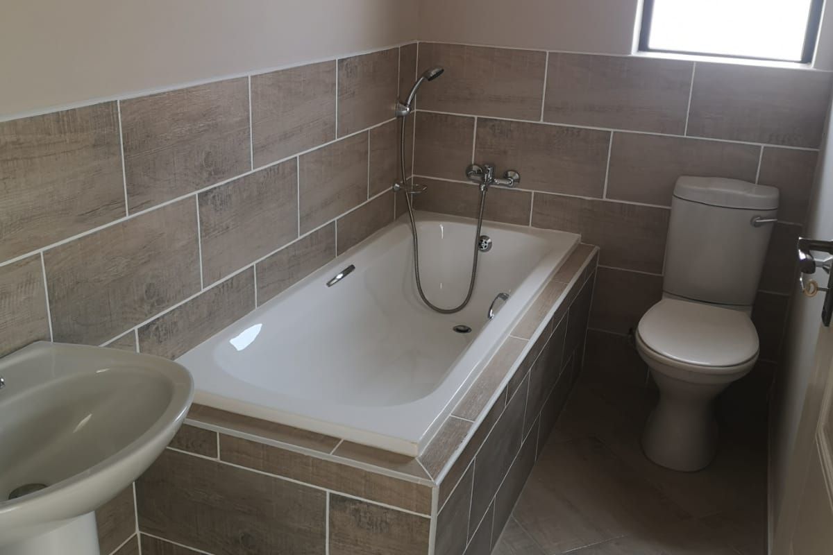A clean, modern bathroom featuring a built-in bathtub with handheld shower mixer, pedestal basin, and toilet, finished with neutral wood-look wall and floor tiles.