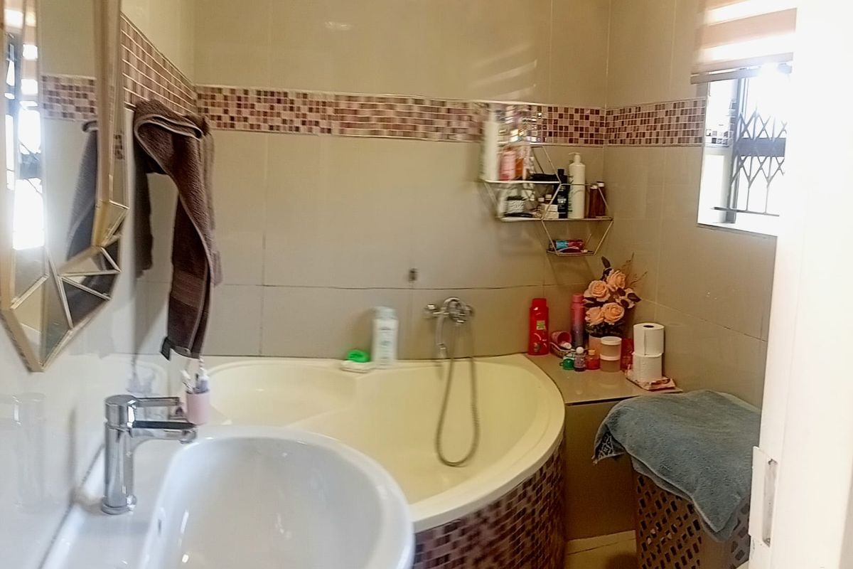A compact bathroom featuring a corner bathtub with mosaic tiling, a pedestal basin, and neutral wall tiles with a decorative mosaic border.