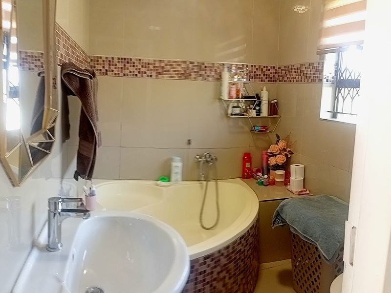 A compact bathroom featuring a corner bathtub with mosaic tiling, a pedestal basin, and neutral wall tiles with a decorative mosaic border.