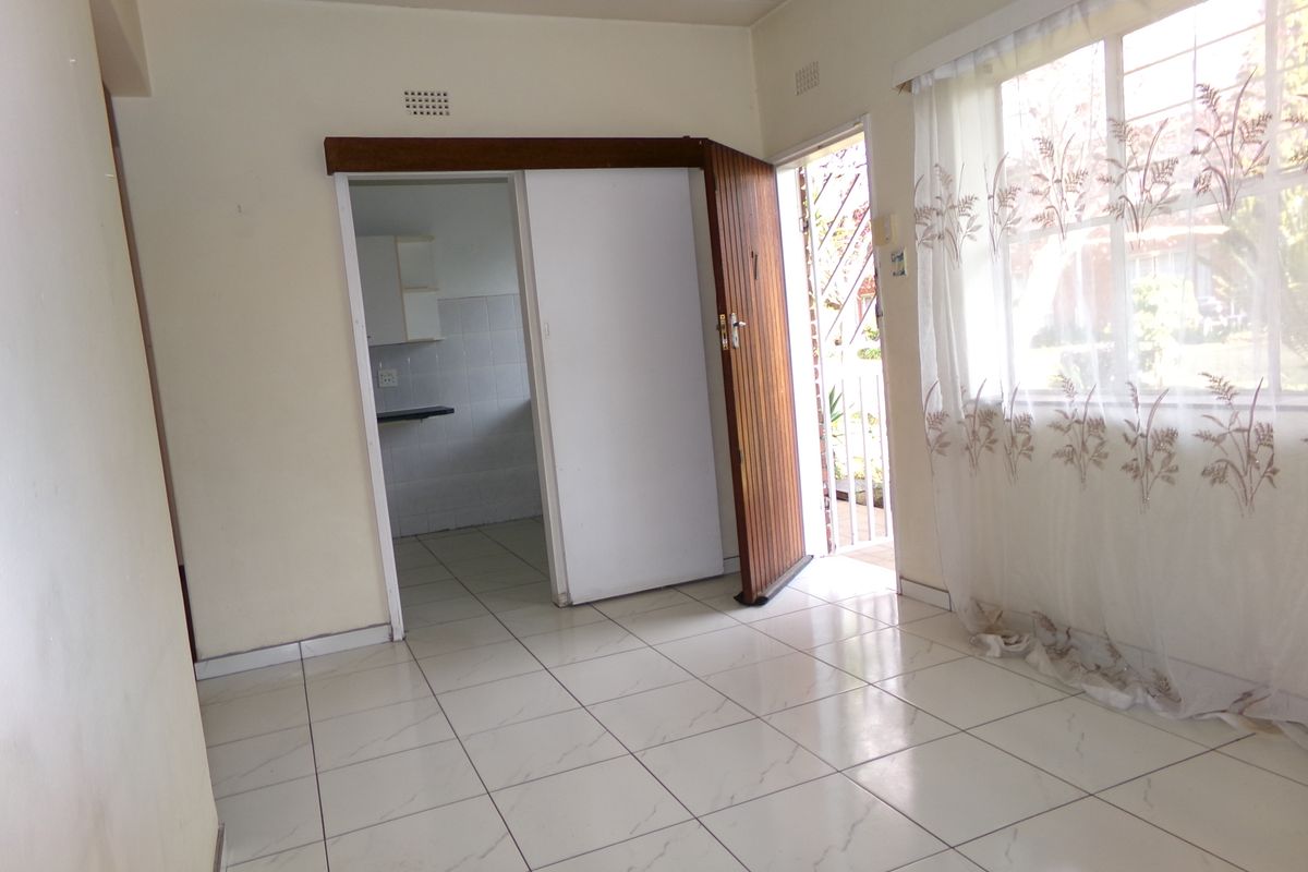 An empty living area featuring tiled flooring, a sliding door, and a window with security bars, leading into a separate kitchen space.