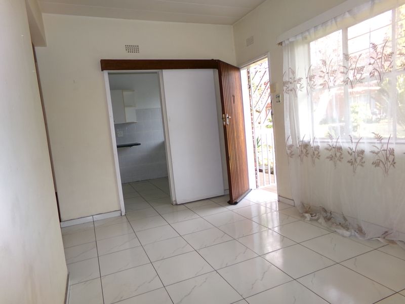 An empty living area featuring tiled flooring, a sliding door, and a window with security bars, leading into a separate kitchen space.