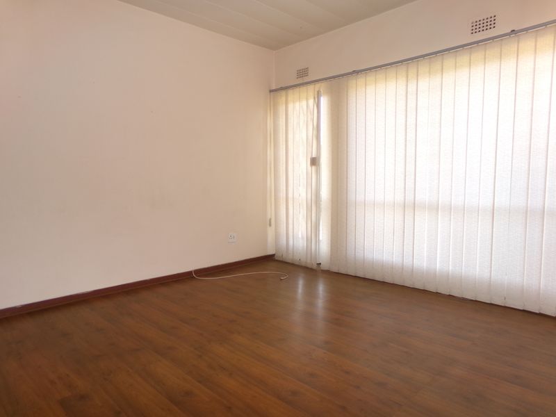 An empty living room featuring warm-toned laminate flooring, neutral walls, and large vertical blinds covering the window area.