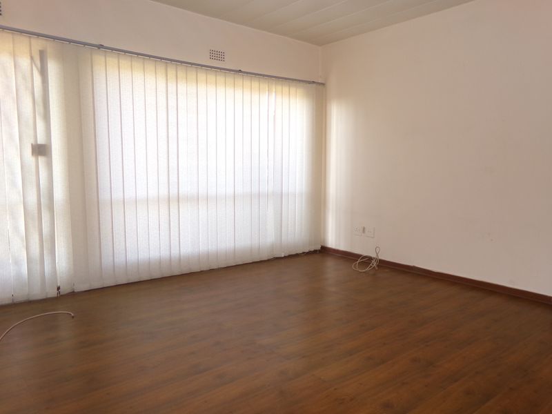 An empty living room featuring laminate wood flooring, neutral walls, and vertical blinds covering a large window.